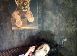 una donna sdraiata a terra morta con in mano una cornetta telefonica rossa e affiancara da un quadro di un leoncino